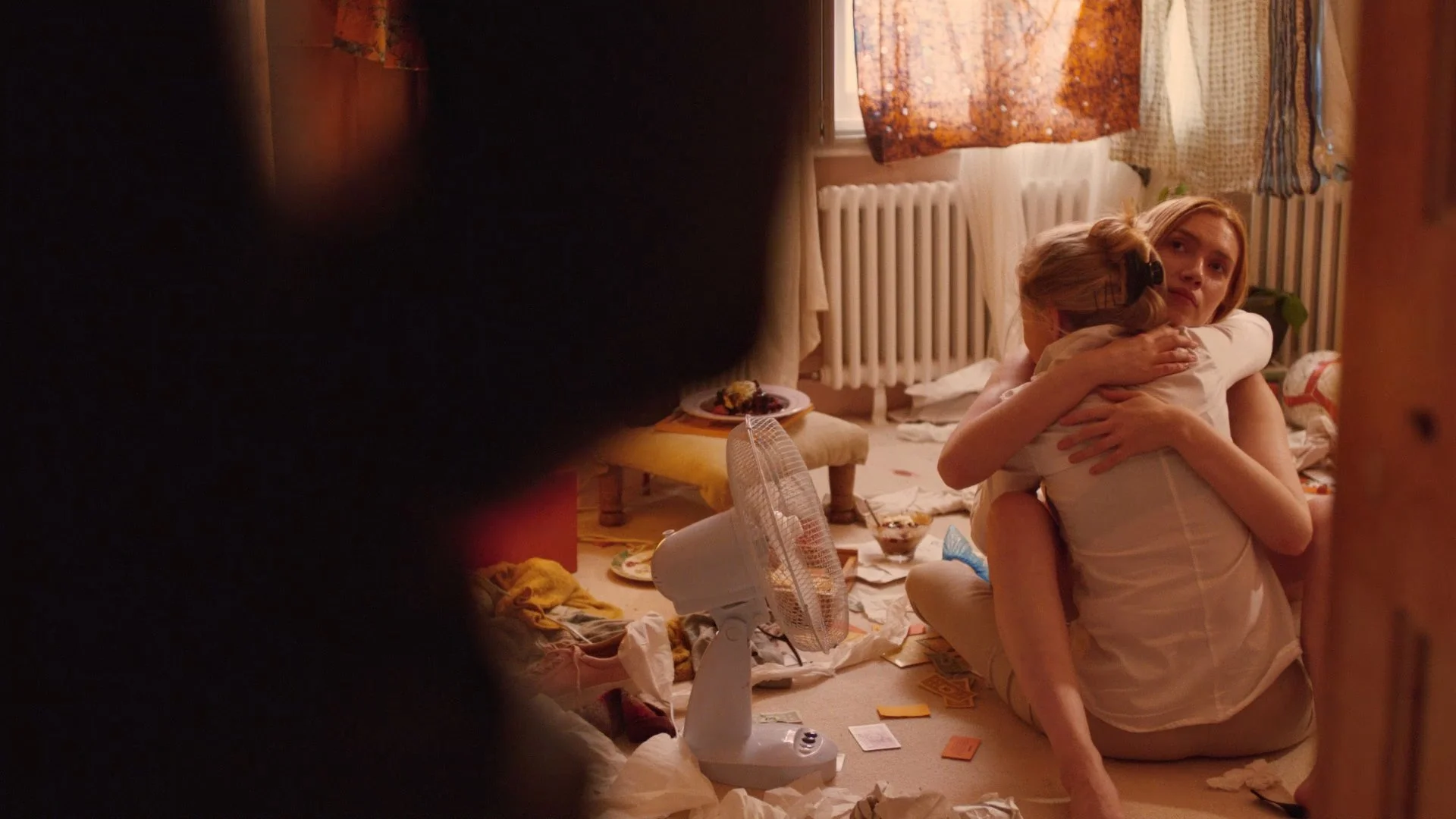 A woman and a young girl are hugging on the floor of a cluttered room, with a window, curtains, and a radiator in the background. There are scattered papers, a fan, and plates with food around them.