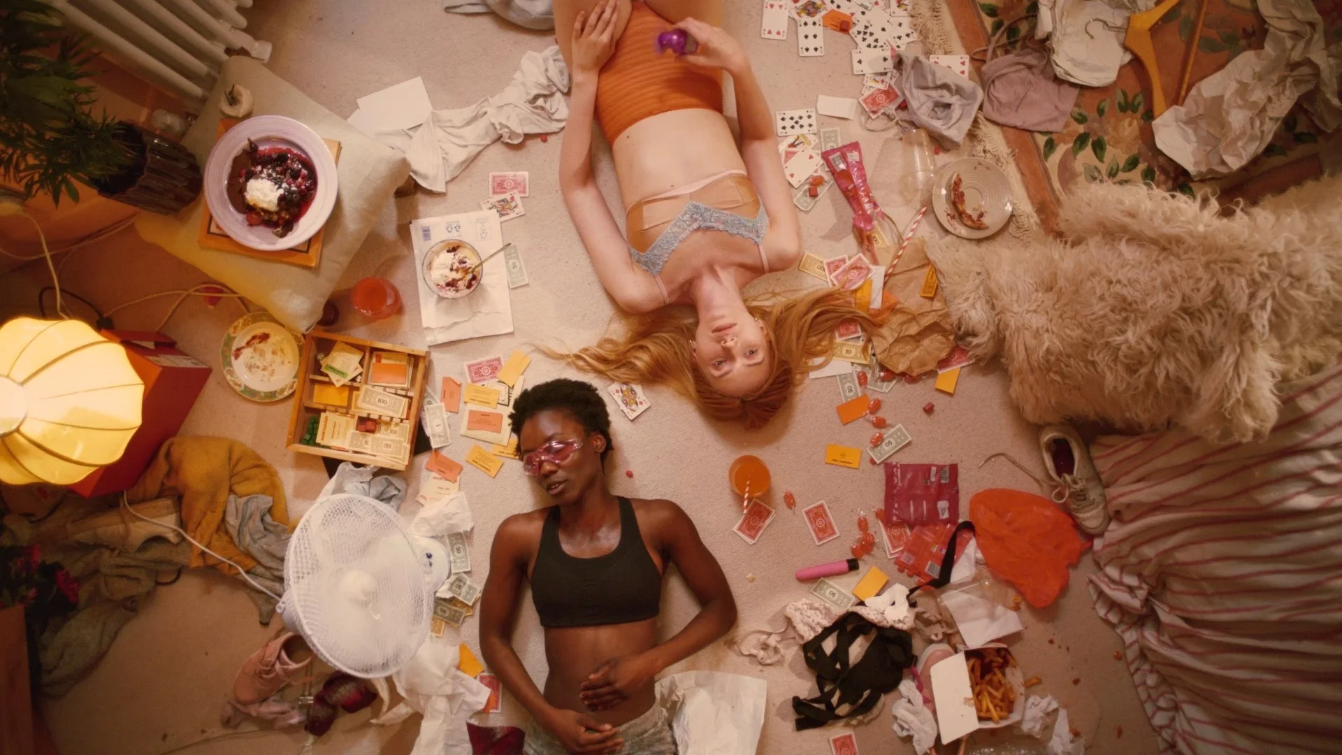 Three women and a dog lying on a cluttered floor after a party, surrounded by scattered snacks, drinks, discarded napkins, and various trash.