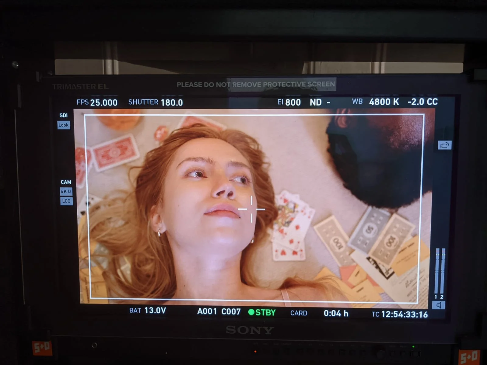 On a camera monitor, a red-haired woman lies on her back surrounded by playing cards and money, looking upward with a neutral expression.