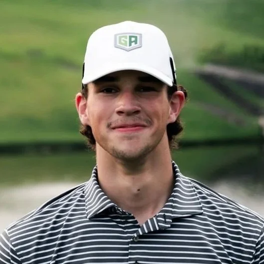 Young man outdoors wearing a white cap with a green logo, striped polo shirt, smiling with a blurred green landscape in the background.