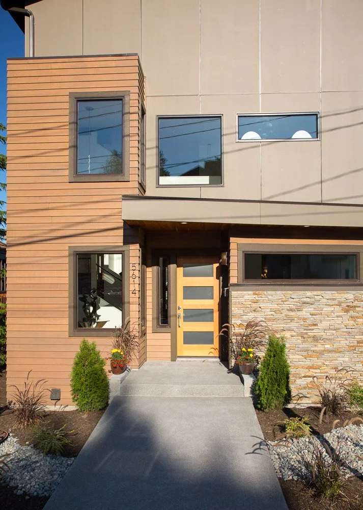 Modern two-story house with a mix of wood, stone, and stucco exterior, featuring a concrete walkway, potted plants, and large windows.