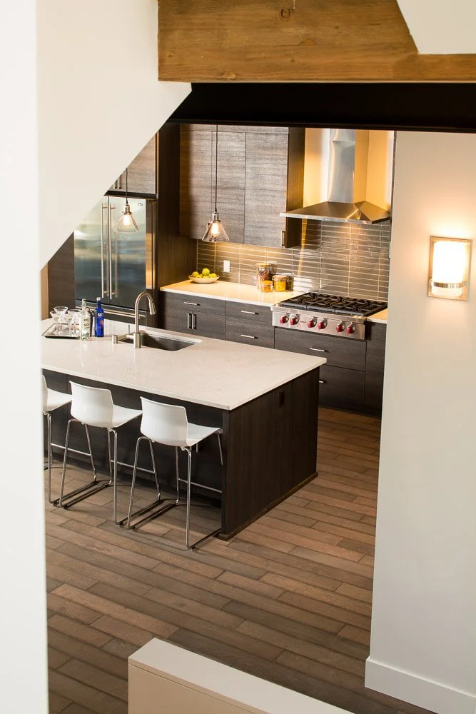 Modern kitchen with dark cabinetry, a white island counter with bar stools, stainless steel appliances, pendant lights, wood flooring, and a brick backsplash.