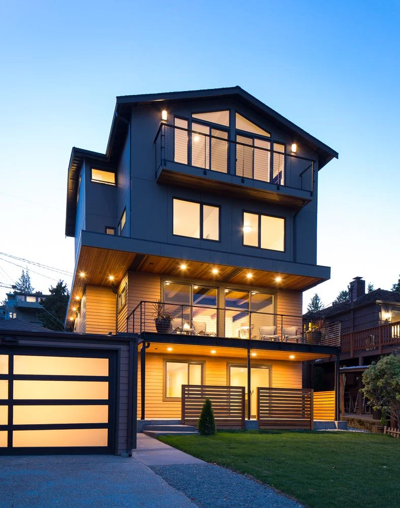 Modern multi-story house with large windows and balconies, illuminated at dusk, front yard with small lawn and fence, neighborhood houses visible.