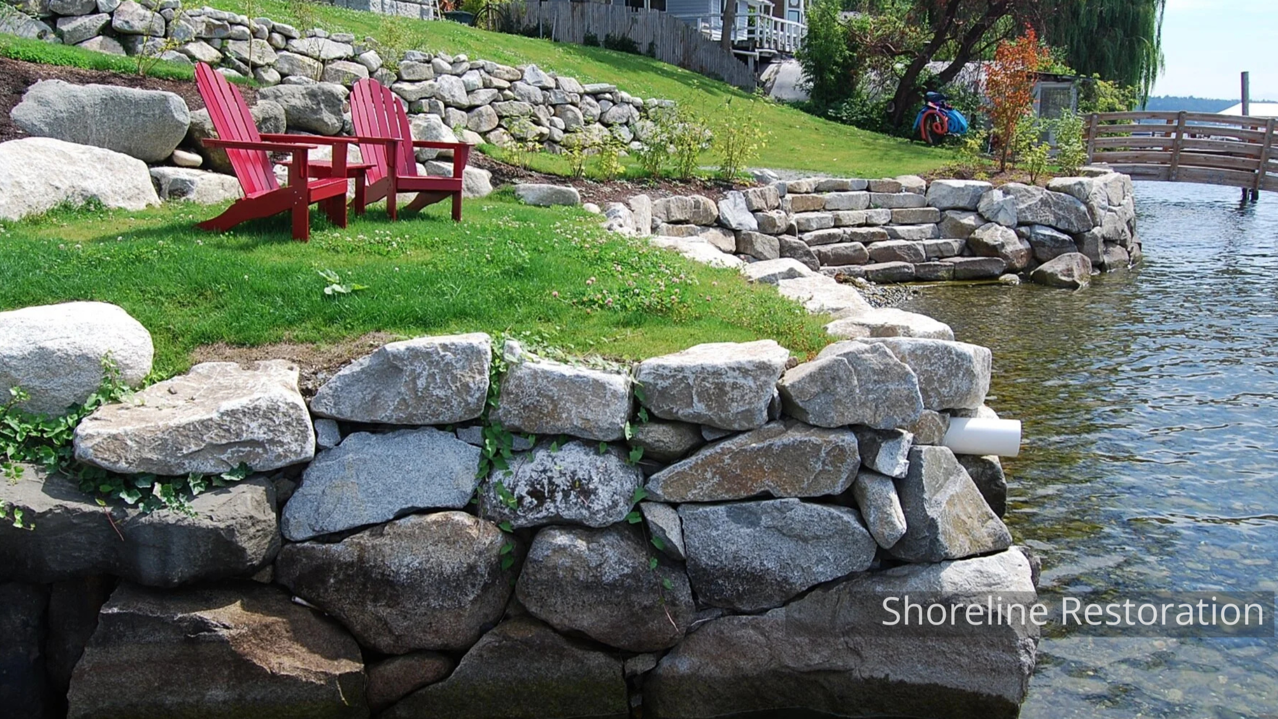 A lakeside backyard with two red Adirondack chairs on a grassy area, a stone retaining wall, and a small wooden bridge over the water. There are trees, plants, and a bicycle in the background.