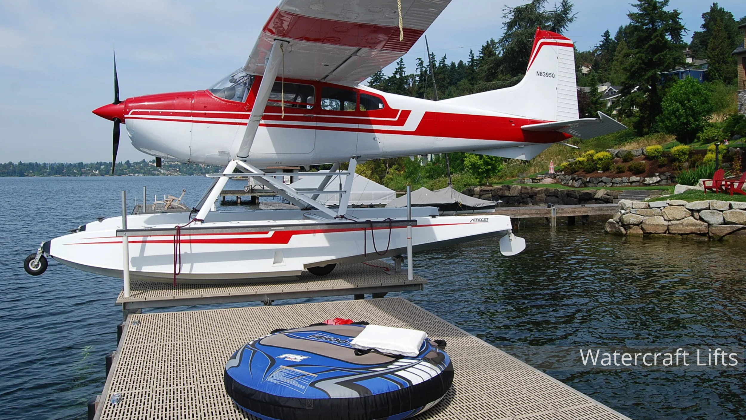 A red, white, and gray seaplane docked on a waterfront with a platform in the foreground. The seaplane is on water, with trees and houses in the background, and a small dock nearby. The platform has a watercraft, a life ring, and some equipment on it