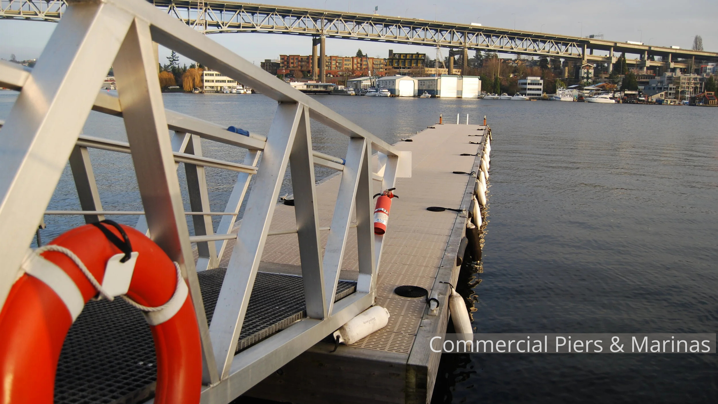 View of a floating dock extending into a body of water with a safety ring attached, and a cityscape with multiple boats, buildings, and a bridge in the background.