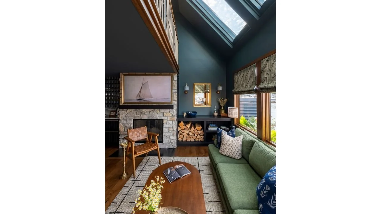 The Nesika Chuck living room pairs deep teal walls with a stone fireplace, warm wood details, and a large overhead skylight. Comfortable seating and windows toward the water create an intimate gathering space filled with natural light.