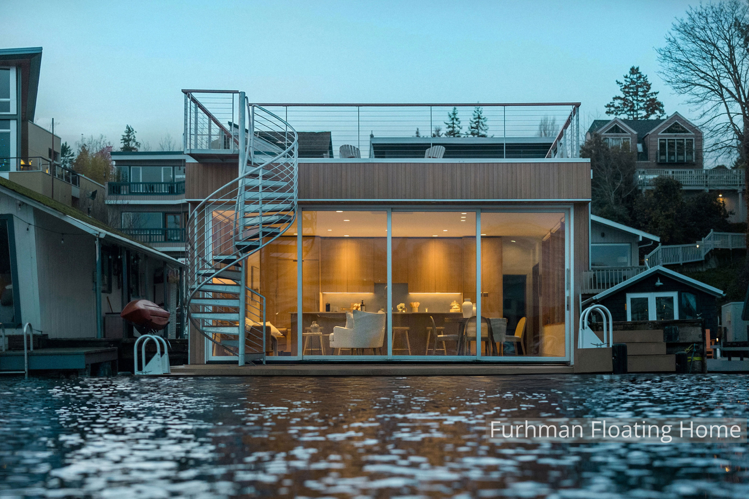 Contemporary floating home with floor-to-ceiling glass, warm wood interiors, and a rooftop deck connected by an exterior spiral staircase. Designed to maximize natural light and waterfront views, the open living and dining space creates a seamless co