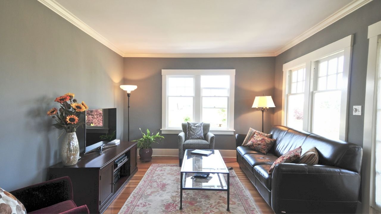 The Greenwood Craftsman living room features warm wood flooring, large windows, gray walls, and a comfortable arrangement of leather seating. Traditional trim and abundant daylight create an inviting space for everyday gathering.
