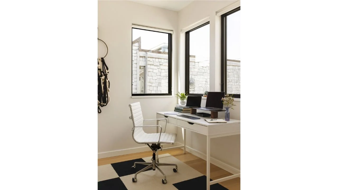 The Mallard Cove home office is positioned beside corner windows that bring daylight and views into the workspace. A streamlined desk and neutral finishes create a bright, practical setting for working within the compact floating home.