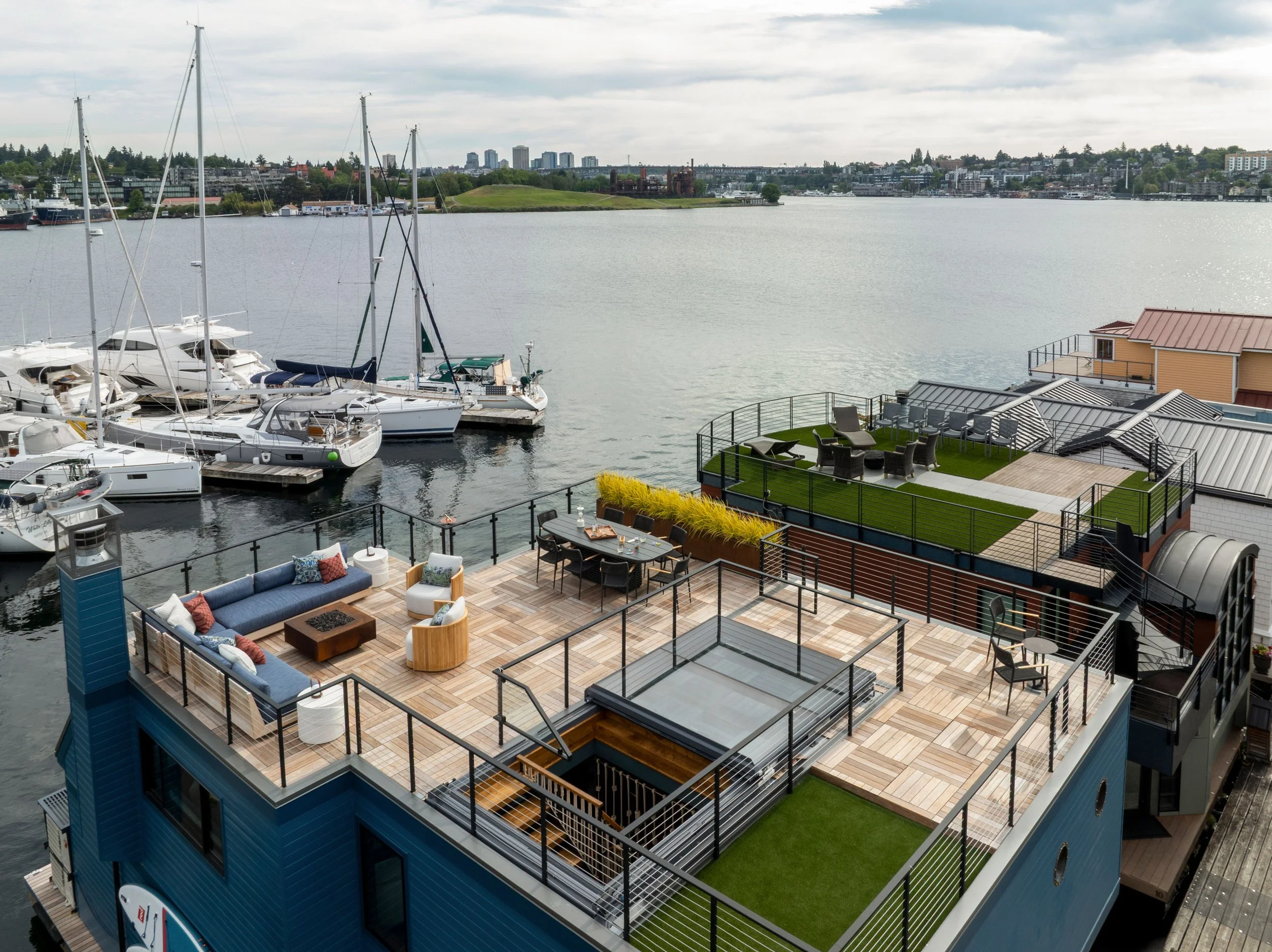 Aerial view of a modern rooftop terrace with outdoor seating, dining area, and lawn, overlooking a marina with yachts and a city skyline in the distance.