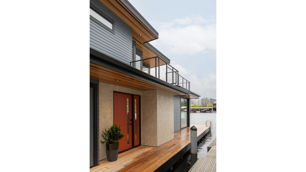 The Shed Buoy’s exterior entry features a bold red front door, warm wood soffits, gray horizontal siding, and a waterfront deck along Portage Bay. Deep roof overhangs and upper-level railings add architectural definition while providing shelter and e