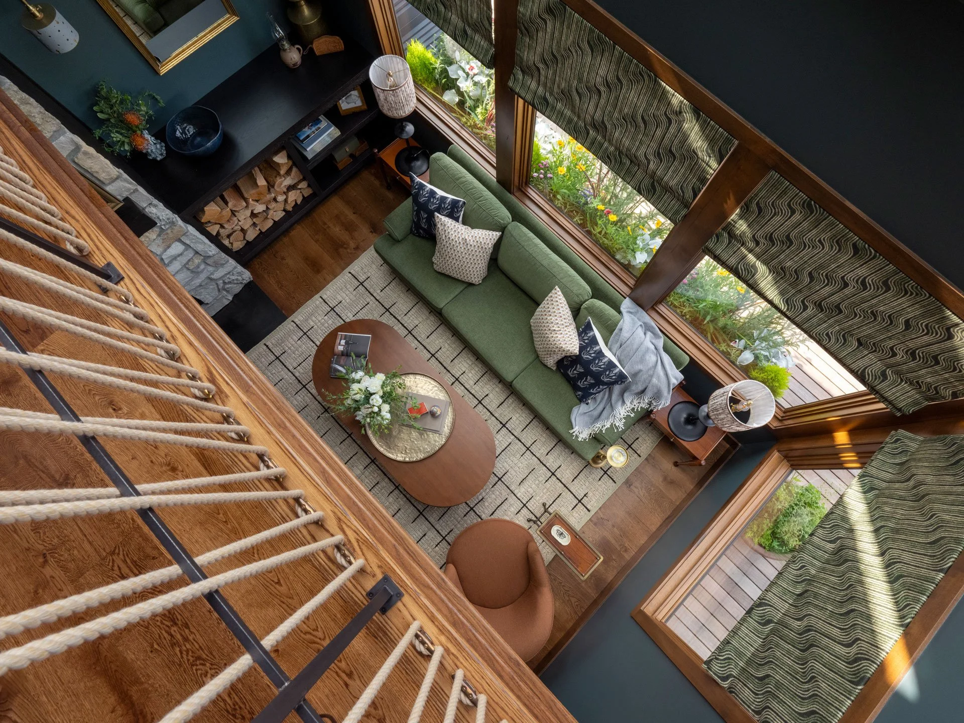 Interior view of a living room taken from a loft space, showing a green sofa with patterned cushions, a wooden coffee table with books and flowers, large windows with green patterned curtains, a bookshelf with firewood, a floor lamp, and decorative i