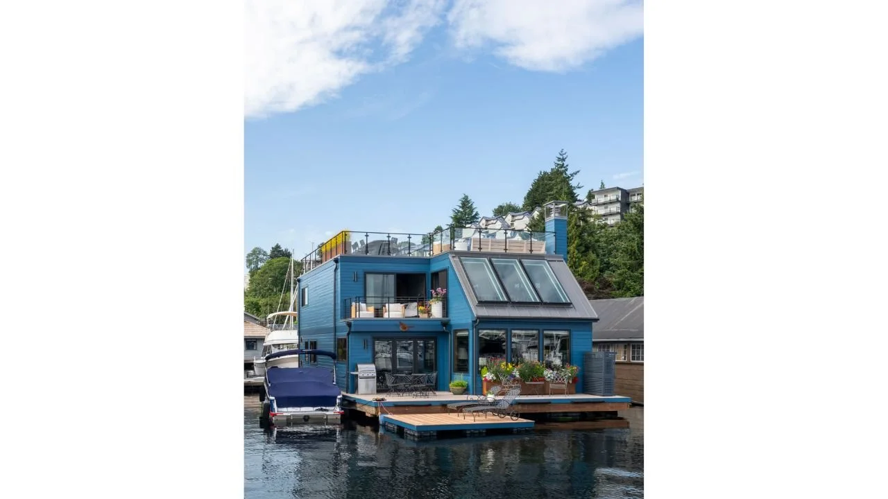 The Nesika Chuck Floating Home features a distinctive blue exterior, expansive waterfront windows, sloped skylights, and outdoor decks positioned directly over the water. The architecture balances a playful residential character with natural light, v