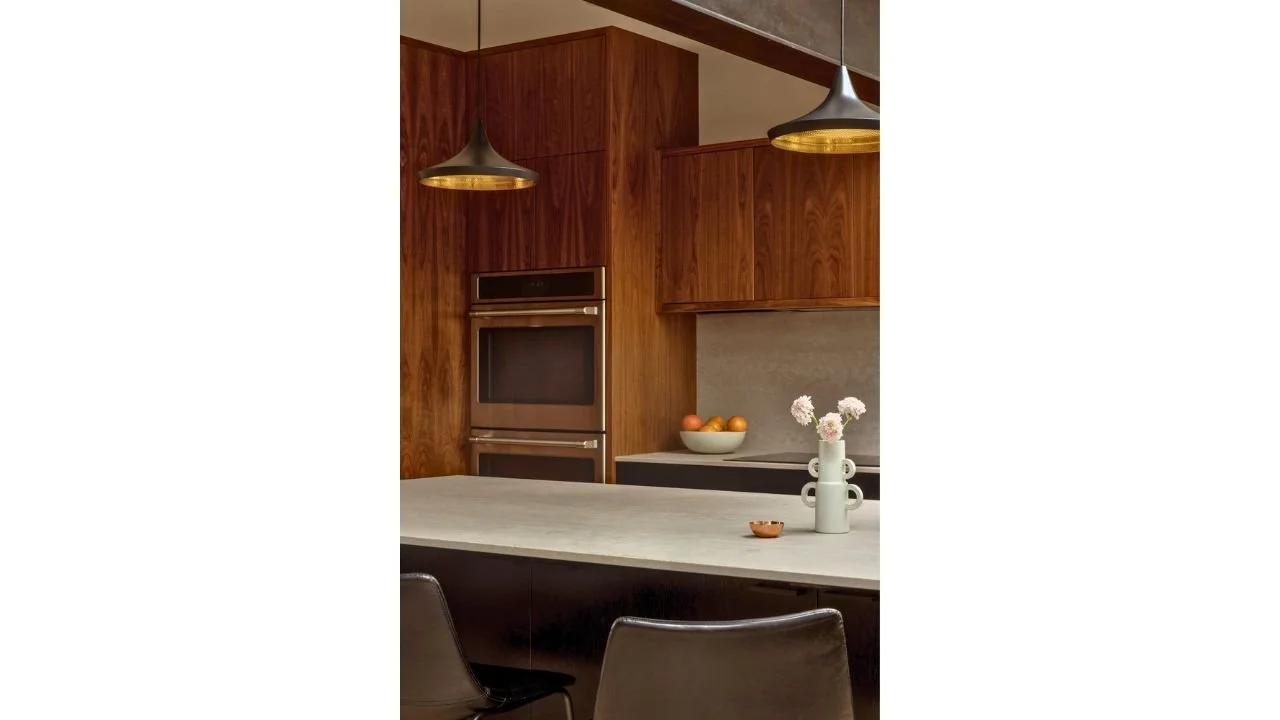 The Mallard Cove kitchen pairs a generous island with warm wood cabinetry and sculptural pendant lighting. Integrated appliances and clean-lined finishes create an efficient gathering space with a polished contemporary feel.