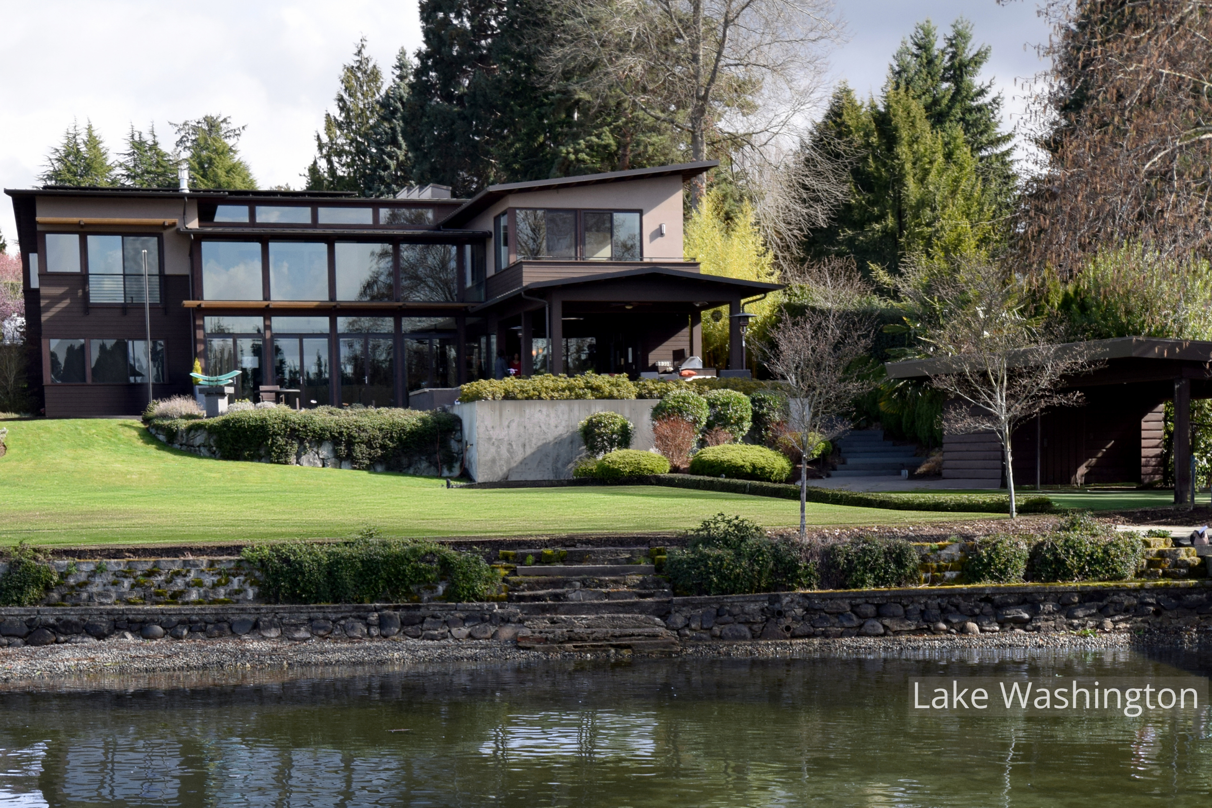 Northwest contemporary home on Lake Washington with expansive floor-to-ceiling windows, layered rooflines, and covered outdoor living spaces overlooking the water. Terraced landscaping, stone retaining walls, and broad lawn areas connect the architec