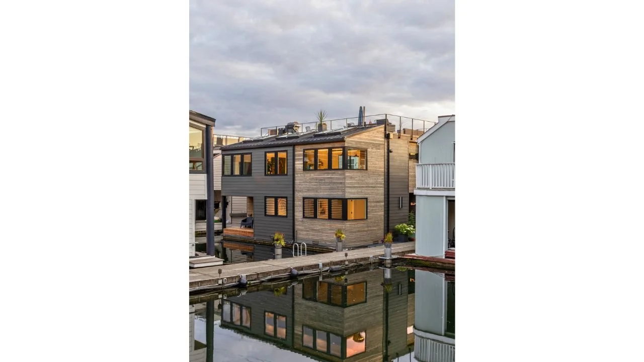 A waterfront view of the Mallard Cove Remodel shows the contemporary floating home reflected in calm water among neighboring residences. Warm interior lighting, broad windows, and layered exterior materials give the remodeled home a distinctive prese