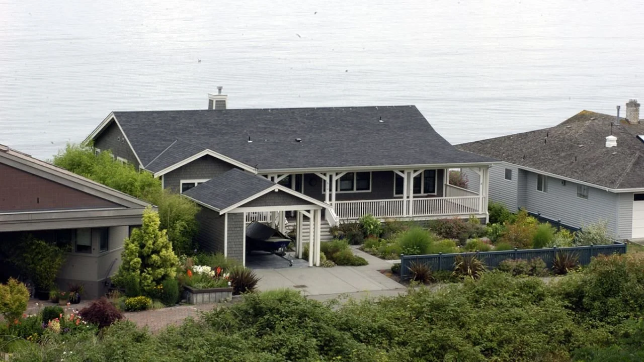 A wide view of the Port Townsend Getaway shows the home, covered entry, landscaped grounds, and surrounding coastal setting. Layered rooflines and sheltered outdoor spaces give the residence a cohesive, welcoming form.