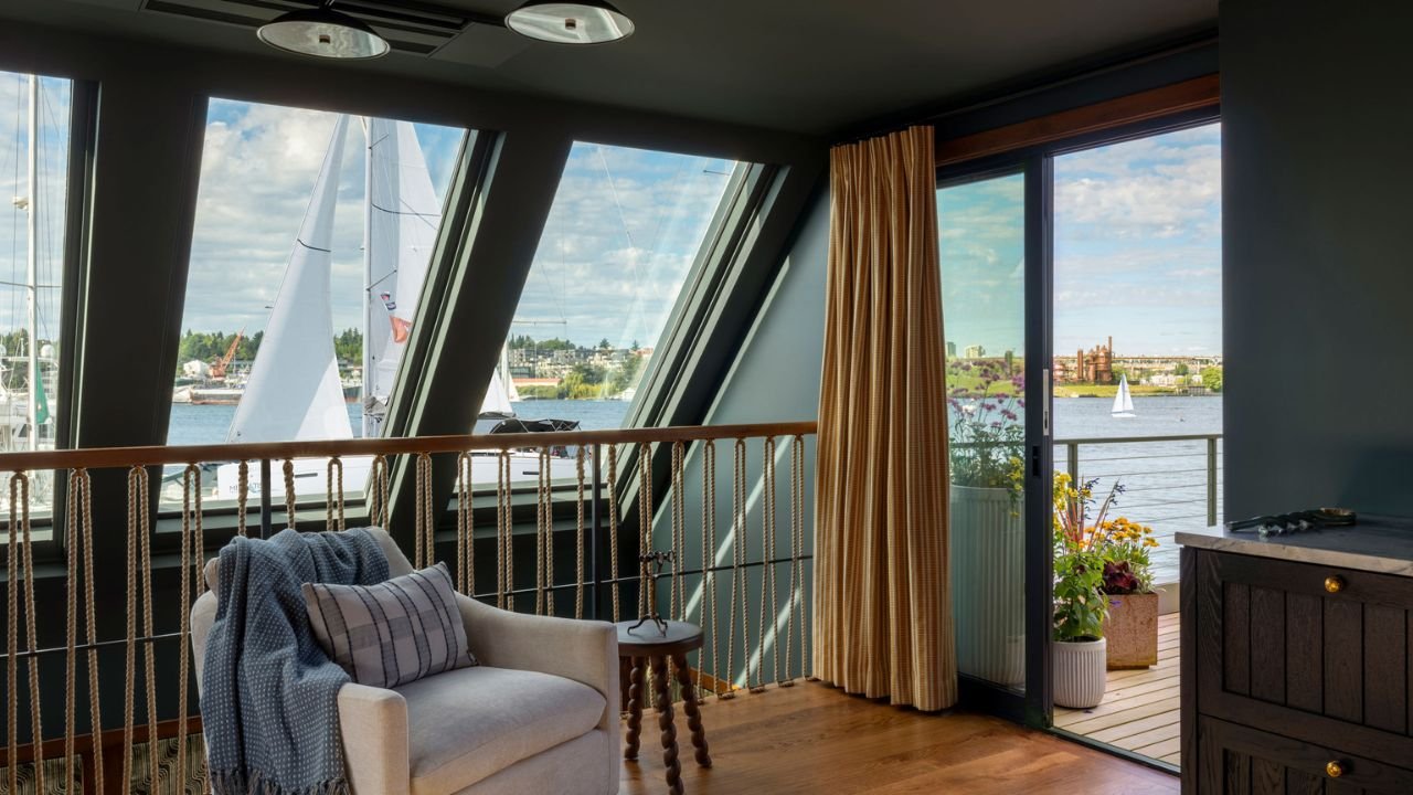 An upper-level reading nook in the Nesika Chuck Floating Home is framed by angled windows and broad views across the water. A comfortable chair, warm wood flooring, and access to the outdoor deck create a quiet place to relax above the main living ar