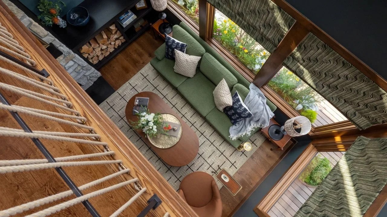 An overhead view from the loft reveals the Nesika Chuck living room arranged around a green sofa, oval coffee table, fireplace, and waterfront windows. The open vertical connection allows natural light to move between levels while making the compact 