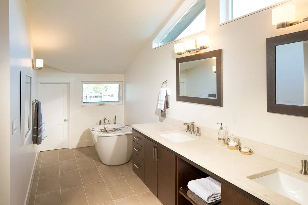 Modern bathroom with double sinks, dark wood vanity, two mirrors, frosted glass lighting, bathtub near window, towel racks, beige floor tiles, and white walls.