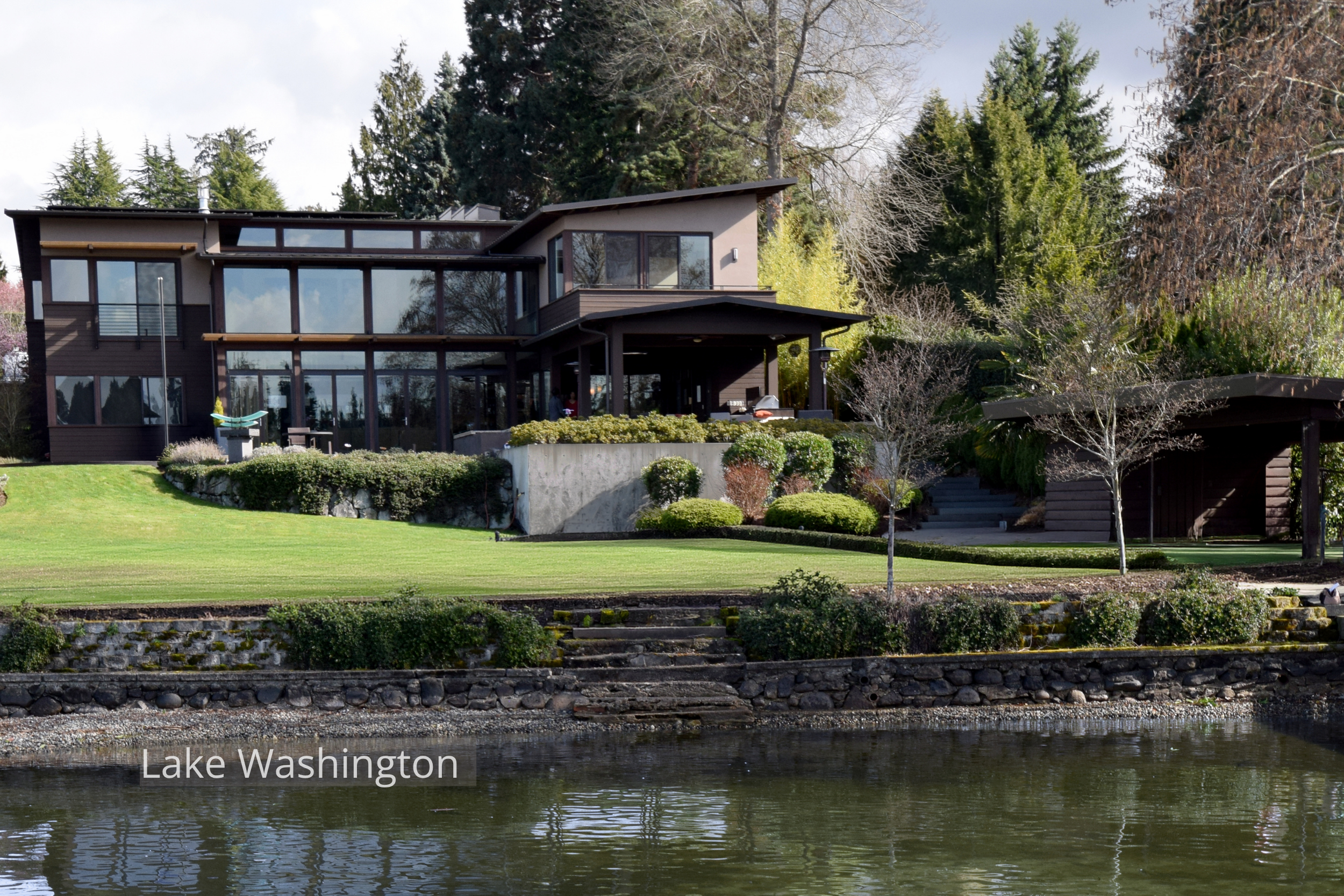 Northwest contemporary home on Lake Washington with expansive floor-to-ceiling windows, layered rooflines, and covered outdoor living spaces overlooking the water. Terraced landscaping, stone retaining walls, and broad lawn areas connect the architec