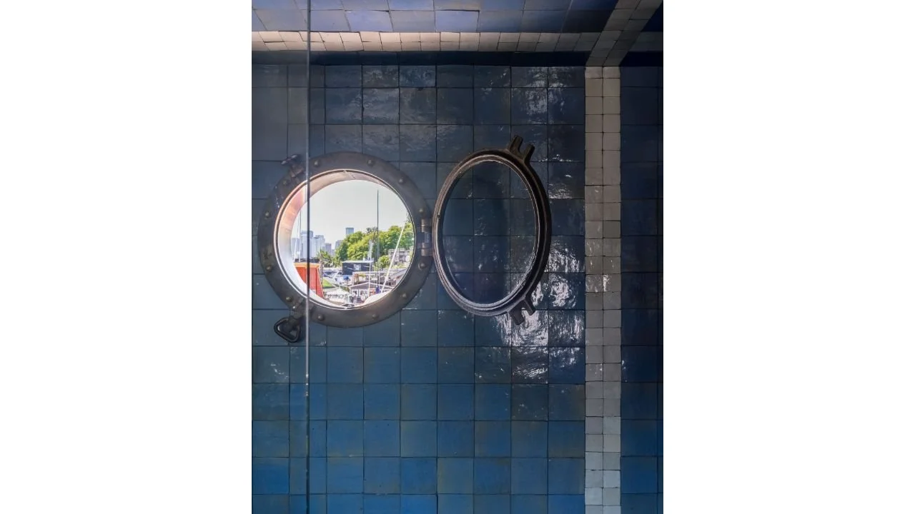 A circular porthole window is set within glossy blue tile at the Nesika Chuck Floating Home, creating a playful nautical connection to the surrounding water. The reflective tile and compact opening bring natural light and character into the interior.