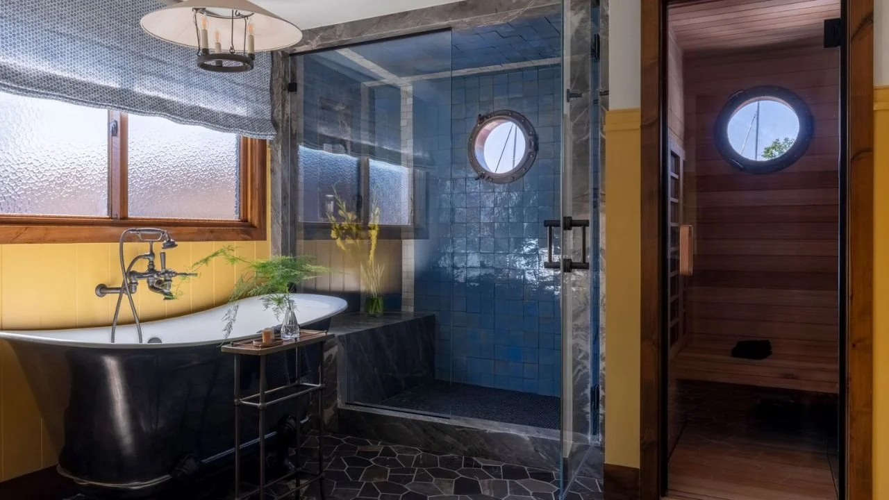 A dramatic bathroom in the Nesika Chuck Floating Home features a dark freestanding tub, glass-enclosed shower, blue tile, and an adjacent wood-lined sauna. Porthole windows, warm lighting, and tactile materials create a private retreat inspired by li