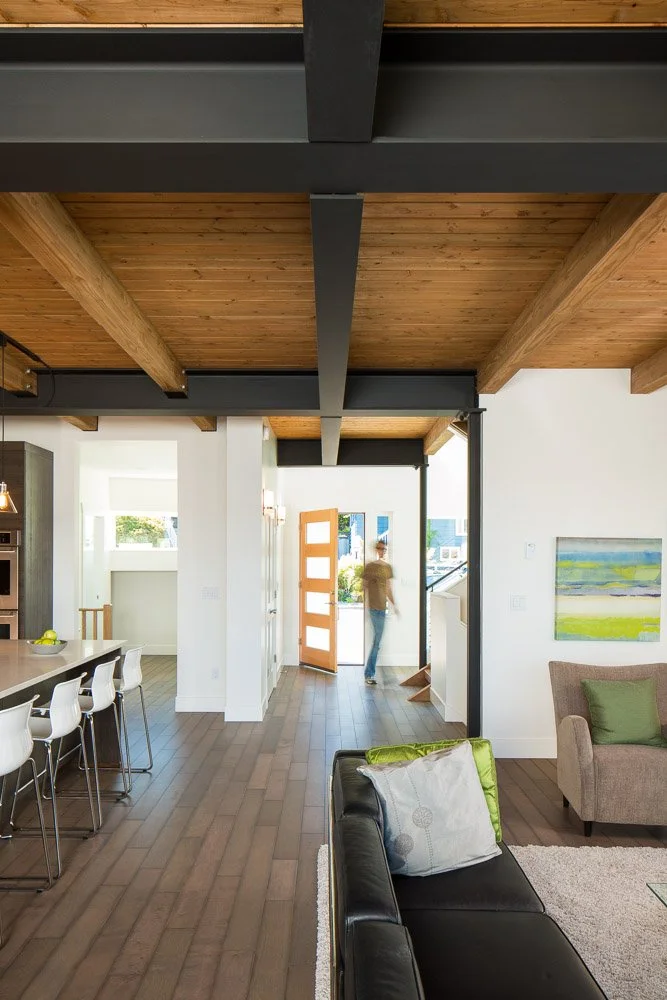 Interior of a modern home with wooden ceiling beams, a black leather sofa with pillows, a beige armchair with a green pillow, a kitchen island with white chairs, and a person walking out the front door.
