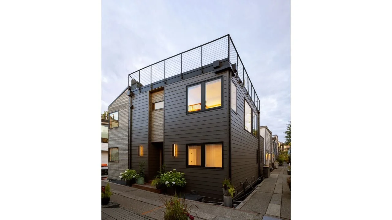 The landward exterior of the Mallard Cove floating home features contemporary gray cladding, warm wood accents, carefully placed windows, and a rooftop terrace. The compact vertical design increases usable living space while maintaining privacy withi