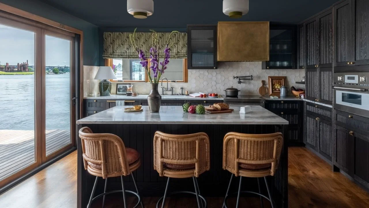 The Nesika Chuck kitchen features dark cabinetry, a generous stone-topped island, woven counter stools, and sliding glass doors overlooking the water. The open layout supports cooking and gathering while maintaining a direct connection to the floatin