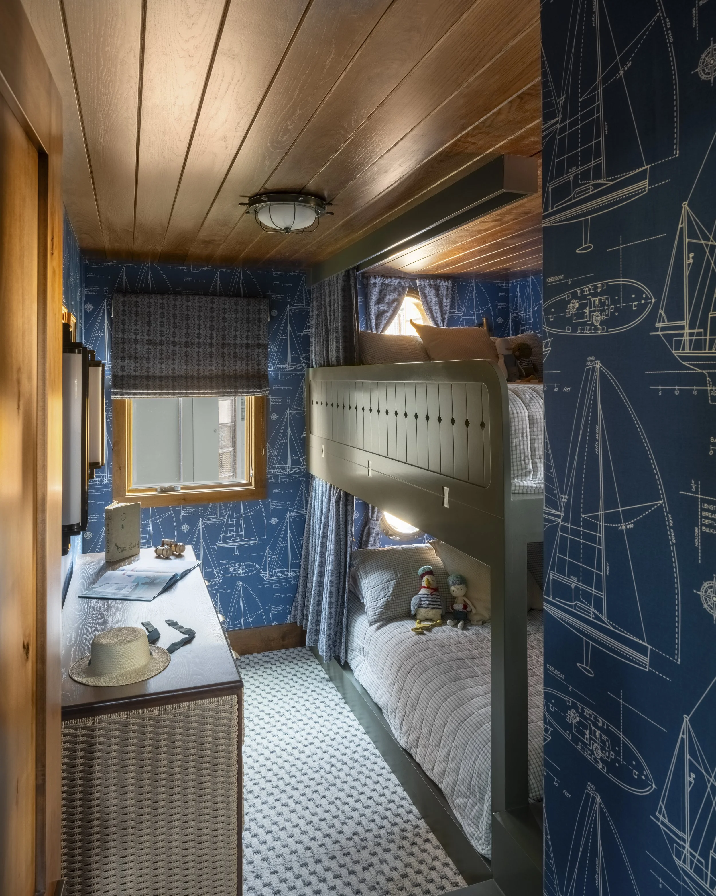 A cozy bedroom with a wooden ceiling and nautical-themed blue wallpaper with white line drawings of ships and boat parts. There is a small window with blue curtains, a bed with a plaid duvet, and stuffed toys. A dresser with decorative items and a ha