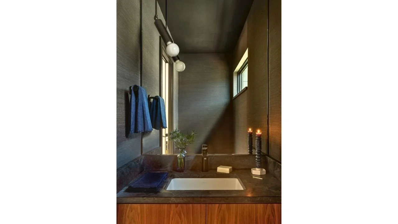 A dramatic bathroom in the Mallard Cove Remodel pairs dark wall tile and countertops with warm wood cabinetry, globe lighting, and a narrow clerestory window. The moody material palette gives the compact space depth, warmth, and character.