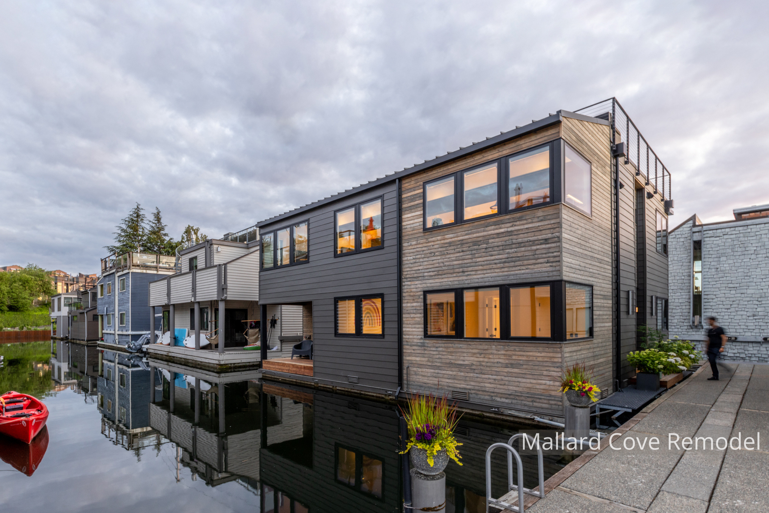 Mallard Cove floating home remodel featuring a refreshed contemporary exterior with natural wood siding, dark horizontal cladding, expansive windows, and a rooftop deck. The updated design brings warmth and modern character to the waterfront residenc