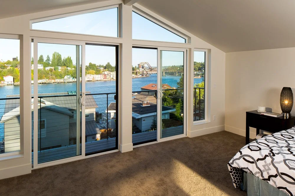 Bedroom with a large window and sliding glass door overlooking a waterway, houses, and a bridge.