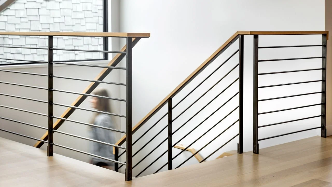 A close view of the Mallard Cove staircase highlights black metal railings, horizontal cables, and a warm wood handrail. The clean detailing allows light to move through the home while adding a refined architectural element to the remodeled interior.