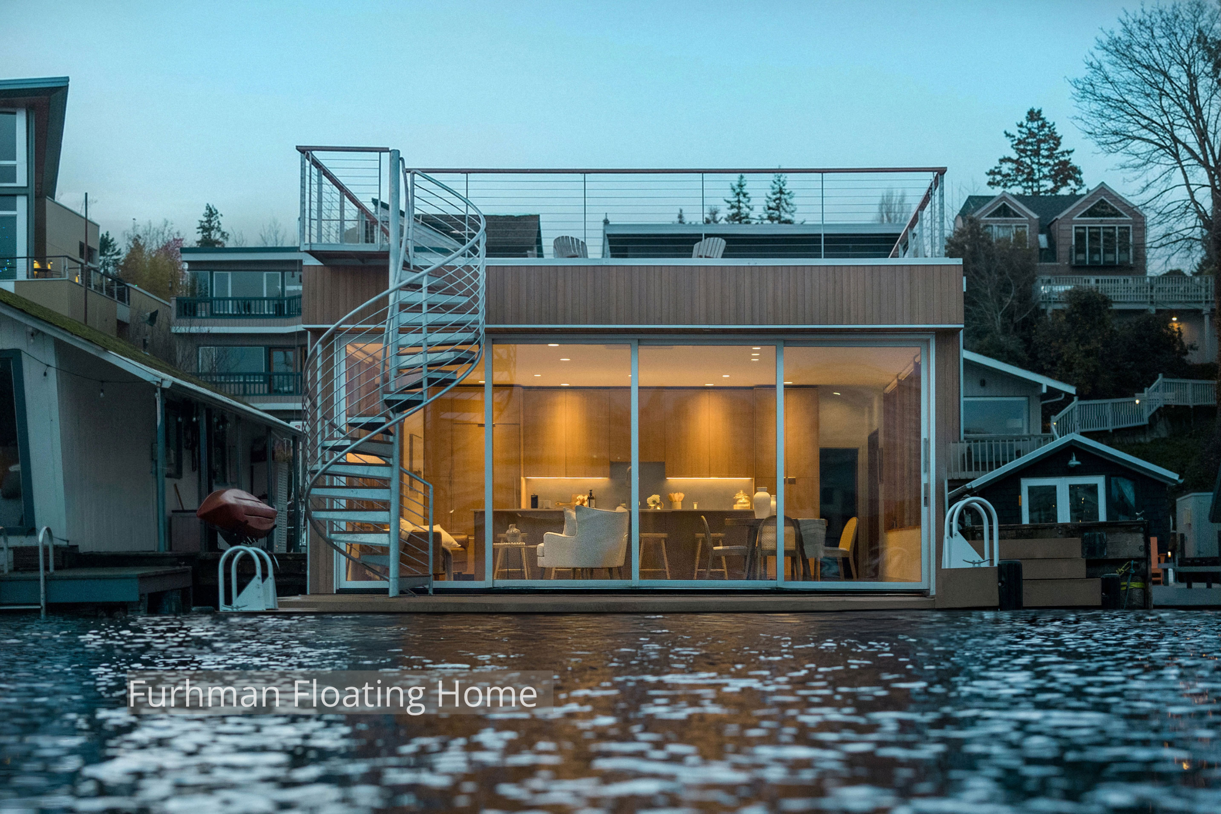 Contemporary floating home with floor-to-ceiling glass, warm wood interiors, and a rooftop deck connected by an exterior spiral staircase. Designed to maximize natural light and waterfront views, the open living and dining space creates a seamless co