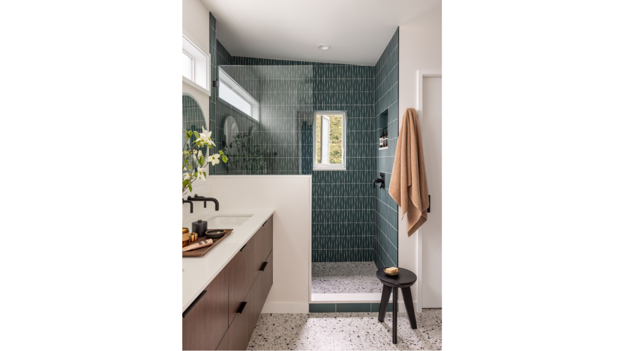 The Shed Buoy’s primary bathroom features deep green vertical tile, a glass-enclosed walk-in shower, terrazzo flooring, and warm wood cabinetry. Matte black fixtures and a narrow window add contrast and natural light, creating a clean, modern retreat