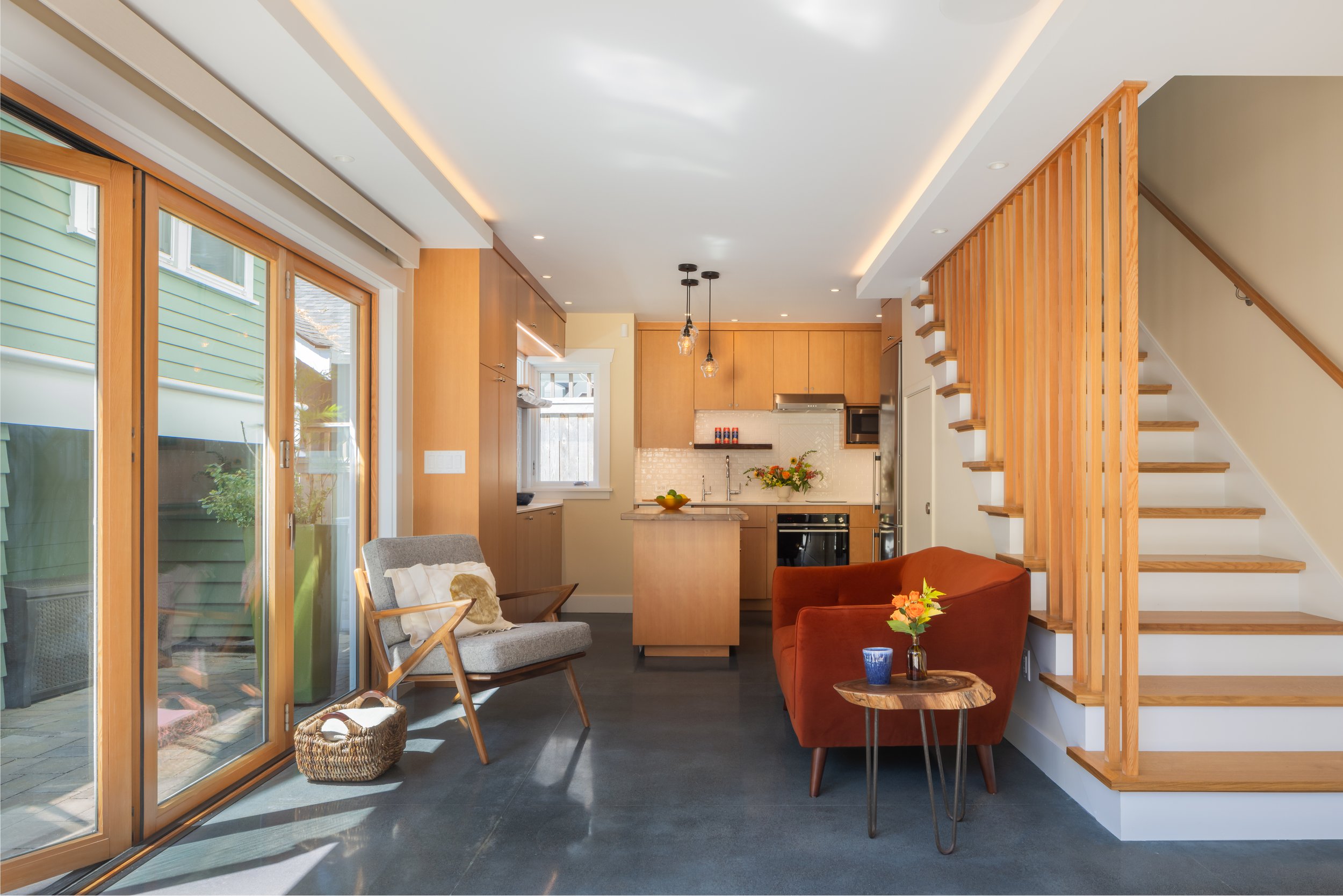 Open-concept interior of a modern Seattle DADU featuring a compact kitchen, living area, and custom wood staircase with vertical slat detailing. Large sliding glass doors bring in natural light and connect the interior to a private backyard patio, em