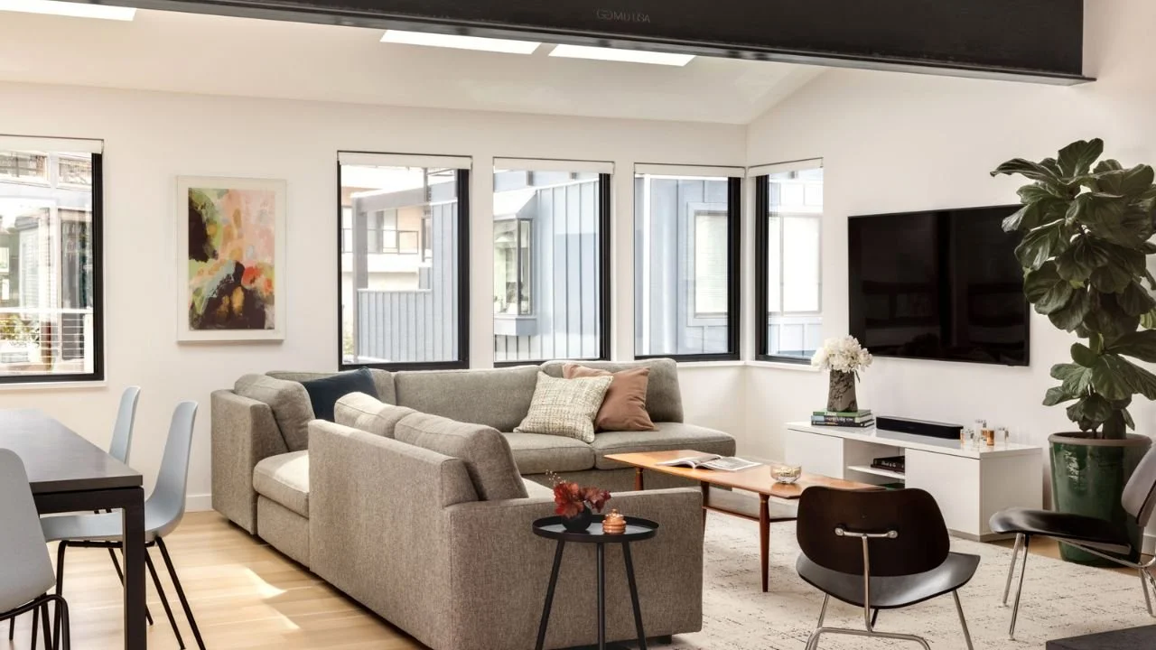 The Mallard Cove living room features comfortable seating, light wood flooring, and windows positioned to capture daylight and waterfront views. The open arrangement creates a relaxed gathering space connected to the adjacent dining and kitchen areas
