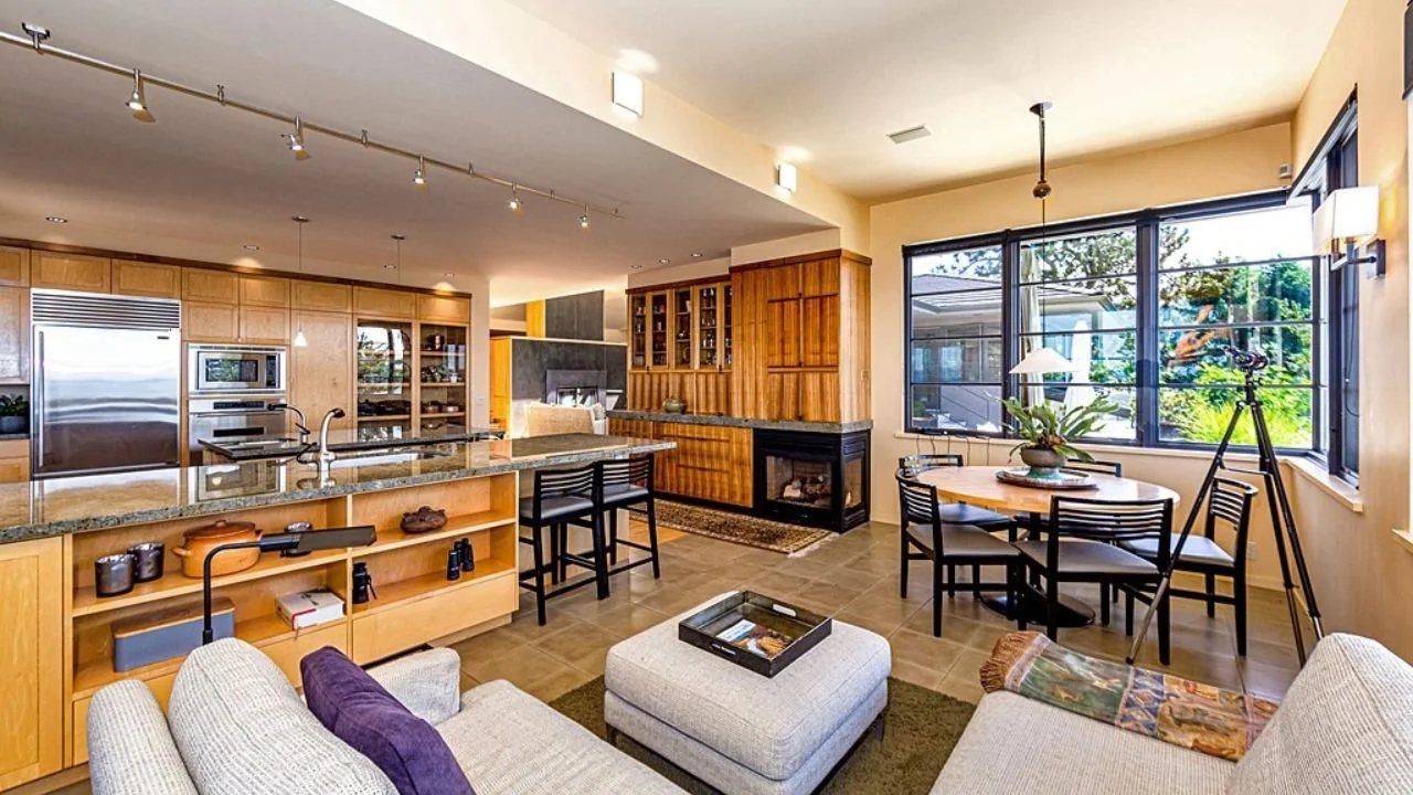 Open-concept living room and kitchen organized around broad waterfront windows and abundant natural light. Warm wood finishes, comfortable seating, and a generous island create a relaxed gathering space designed for life in the San Juan Islands.