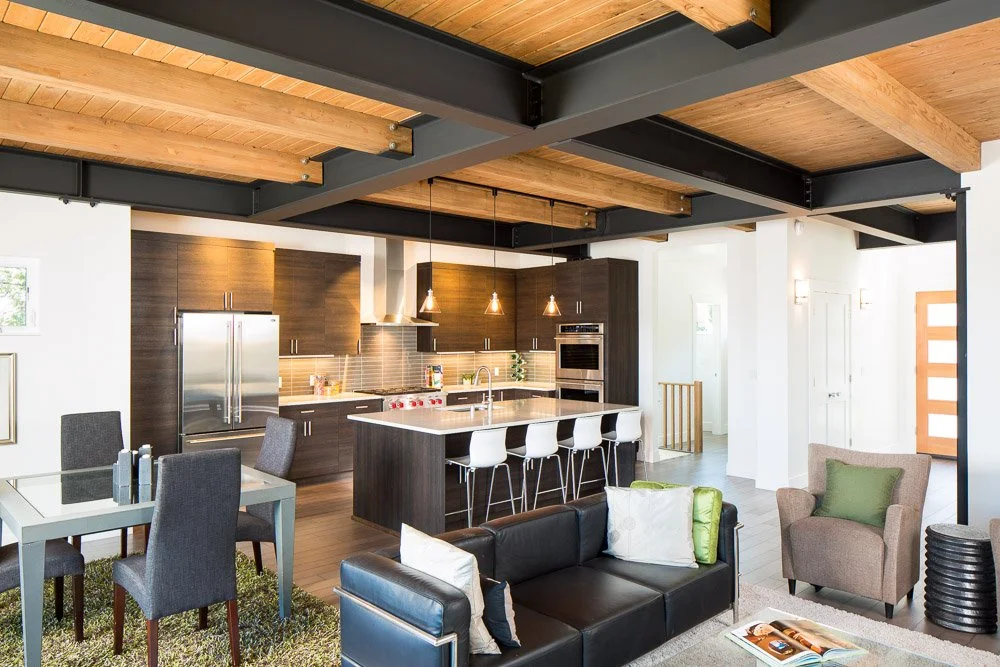 Open-concept living room and kitchen with modern appliances, dark wood cabinetry, and a kitchen island with white bar stools. The dining area has a glass table with dark upholstered chairs, and there are cozy armchairs and a couch with cushions.