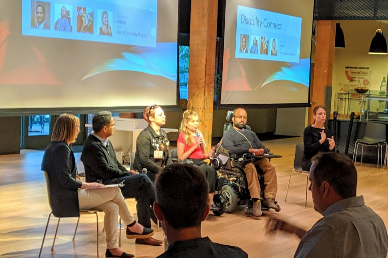 Image Description: Four panelists, a moderator present in front of attendees at Disability Connect, along with an ASL interpreter .