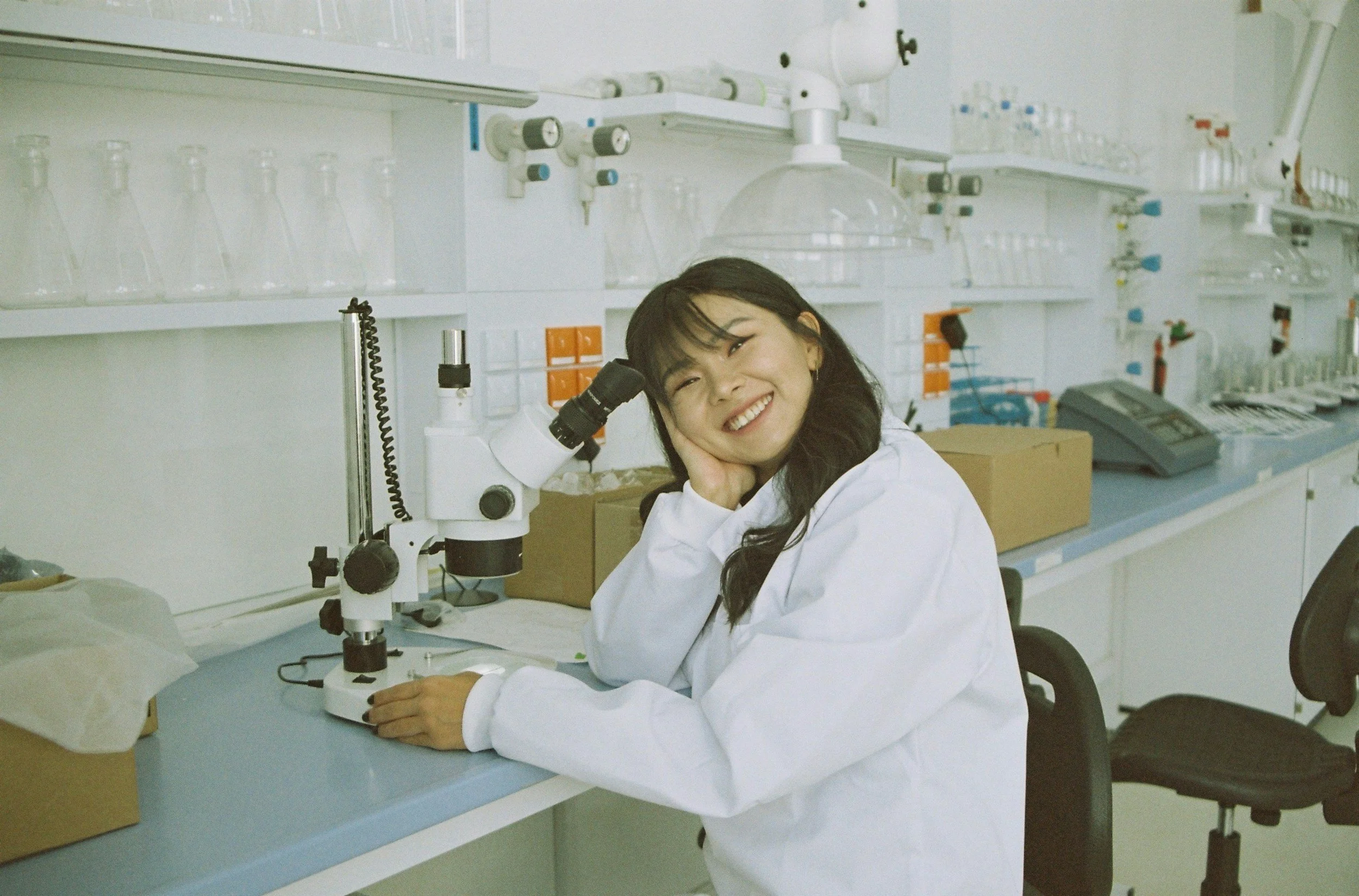 A woman in a laboratory wearing a white lab coat, sitting at a lab counter beside a microscope, smiling and resting her head on her hand.