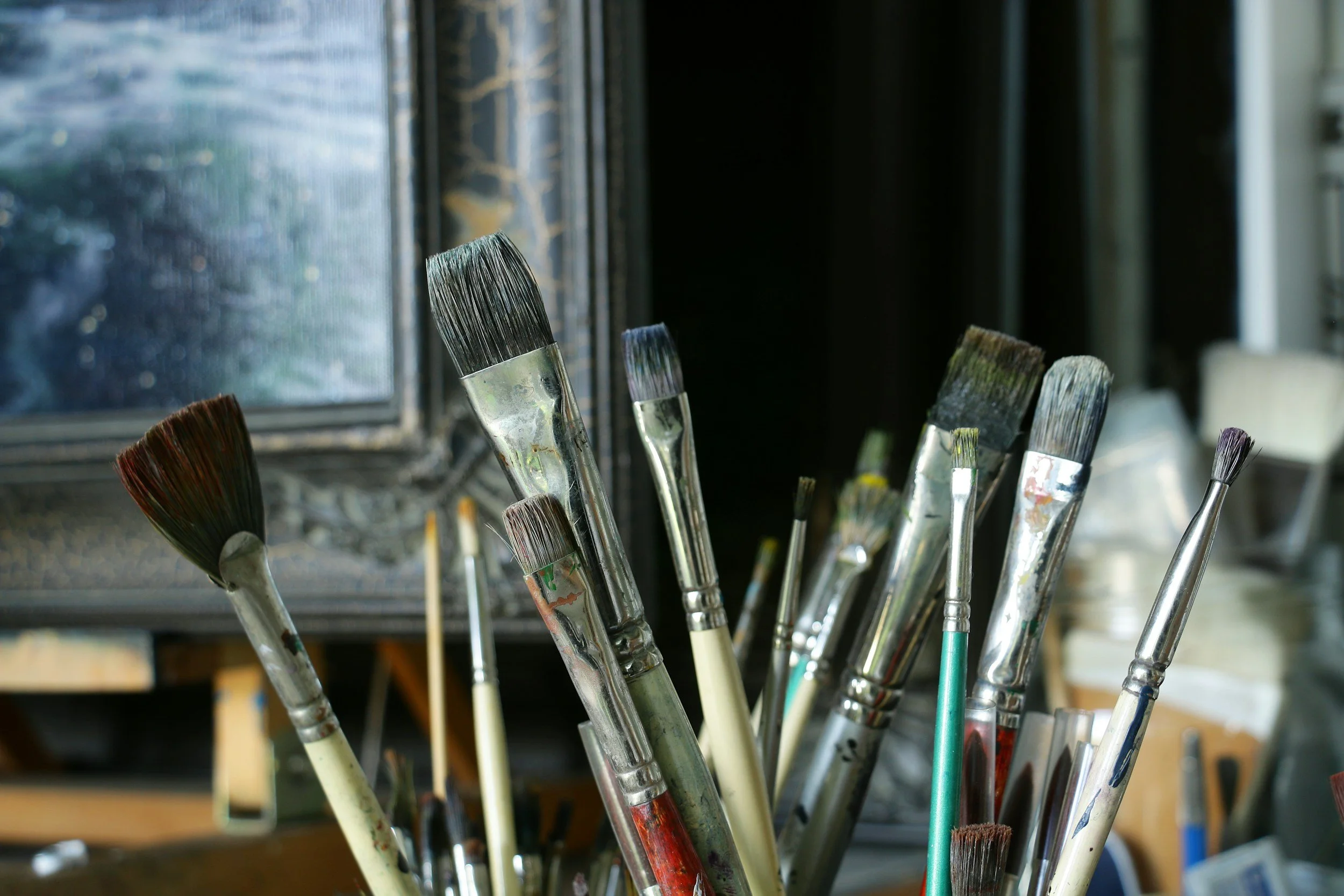 A collection of paintbrushes of various sizes and shapes, some with dried paint on their bristles, standing upright in a container near a window.