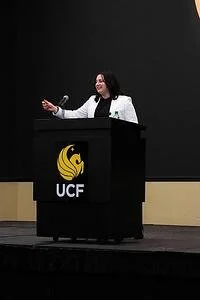 Woman speaking at a podium with UCF logo, in front of a black backdrop.