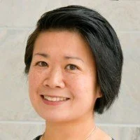 A woman with short black hair smiling, standing against a light-colored tiled wall.