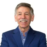 A smiling older man with gray hair and a mustache, wearing a blue suit jacket and a blue dress shirt, against a white background.