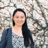 A young woman with long black hair smiling, standing outdoors in front of a wall with vines and red berries.