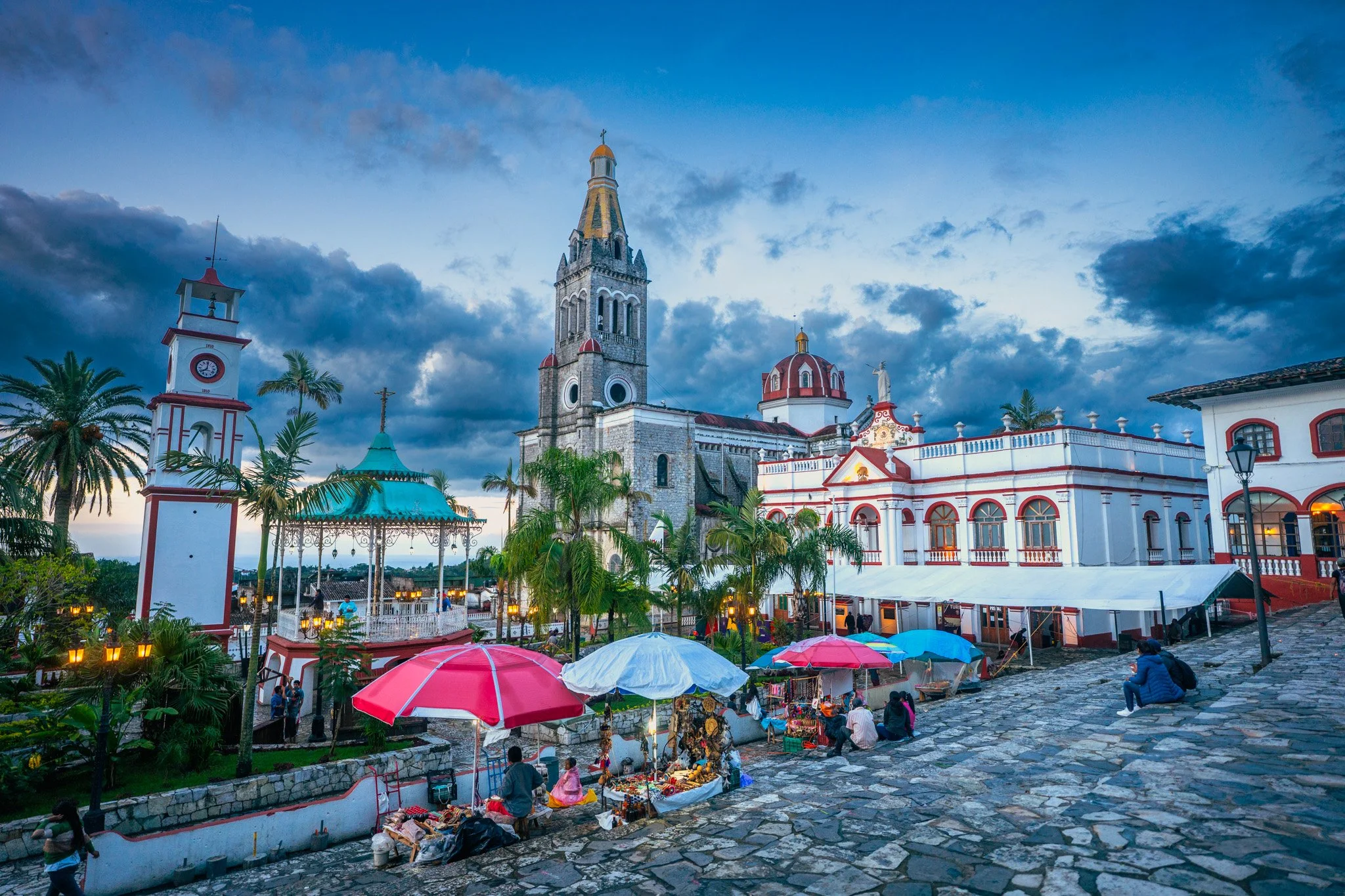 A colorful outdoor market scene in front of a historic church with a tall clock tower, surrounded by palm trees, street vendors with umbrellas, and tourists sitting on cobblestone pavement at sunset.